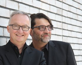 Die beiden Kabarettisten Peter Schaal-Ahlers und Søren Schwesig stehen vor einer gemauerten, weißen Wand. 