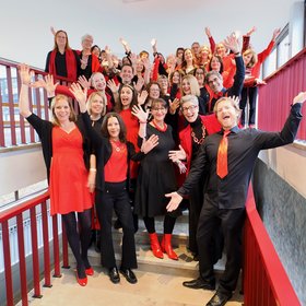 Cruise of Music Rot-schwarz gekleidete Chormitglieder stehen lächelnd und posierend auf einer Treppe.