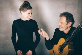 Ein Mann mit Gitarre redet mit einer Frau, beide schwarz gekleidet, vor einer grauen Wannd