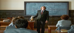 Sitzende Schüler von hinten, auf dem Lehrertische steht ein Pinguin, daneben der Lehrer 
