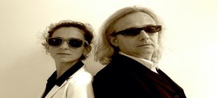 Ein Mann und eine Frau in schwarz-weißer Kleidung mit Sonnenbrille vor einer weißen Wand