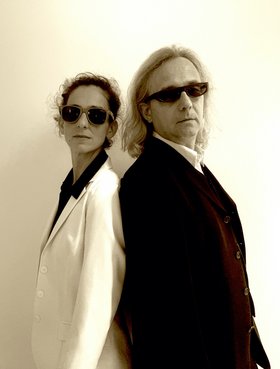 Ein Mann und eine Frau in schwarz-weißer Kleidung mit Sonnenbrille vor einer weißen Wand