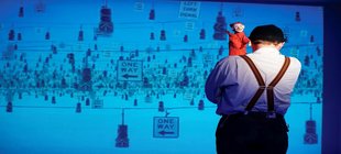 Schauspieler Nobert Eilts steht im heißen Hemd und Hosenträgern vor einer Leinwand mit dem Rücken zum Publikum, über seine Schulter lugt eine Kasperle-Puppe.