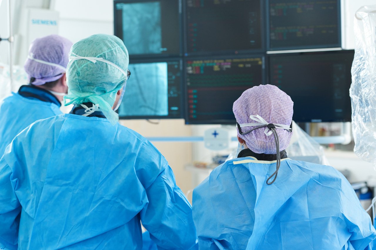 Drei Ärzte vor mehreren Monitoren kontrollieren den Fortschritt beim Einsetzen einer Aortenklappenprothese