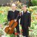 Zwei Musiker in schwarzer, eleganter Kleidung stehen in einem blühenden Garten zwischen roten Tulpen. Eine Person hält ein Cello, beide präsentieren sich in ruhiger, gepflegter Umgebung im Freien.
