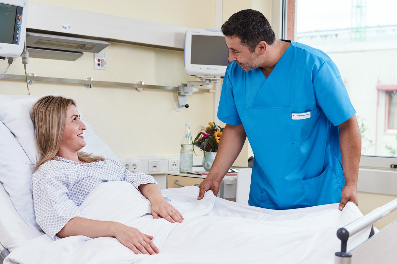 Ein Pfleger wendet sich einer einer im Bett liegenden Patientin zu und spricht mit ihr