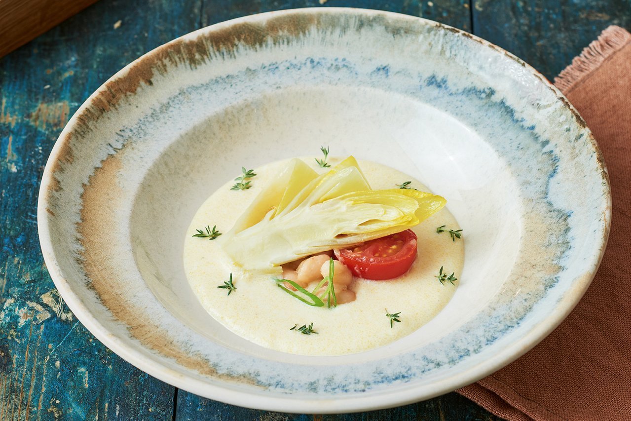 Ein tiefer runder Teller mit blau-braun dekoriertem Rand steht auf einer Shabby-Chic blauen Holzunterlage. Star des Tellers ist eine Chicorée Hälfte, die auf einer hellen zarten Polentacreme liegt. Eine Kirschtomatenhälfte schaut unter dem Salat hervor.