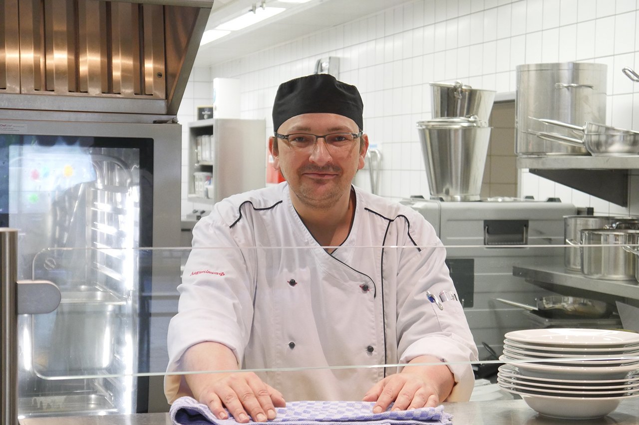 Ein Koch der Augustinum Inkusiven Gastronomie steht hinter einem Tresen zur Essensaugabe und schaut in die Kamera