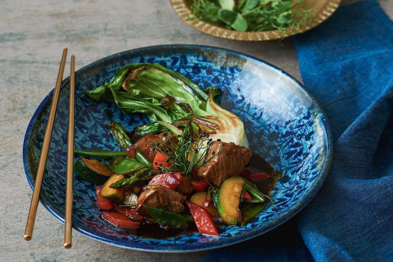 Das Bild ist blau gehalten. Auf einem blauen tiefen Teller mit dunkelblauen Ornamenten ist ein Pfannengericht aus Rindfleisch und buntem Gemüse angerichtet. Auf dem Tellerrand liegen zudem goldene Stäbchen.