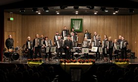 „Mozart, Mancini and more!“ Ein Akkordeon-Orchester mit Dirigent und Schlagzeuger steht auf der beleuchteten Bühne.