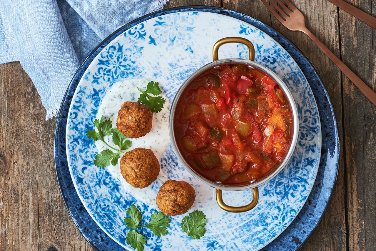 Das Bild ist blau gehalten. Zwei Teller in unterschiedlichen Blautönen mit blauen Ornamenten stehen übereinander. Darauf sind drei Falafel im Halbkreis angerichtet. Daneben steht ein goldenes Töpfchen mit Shakshuka Ragou.