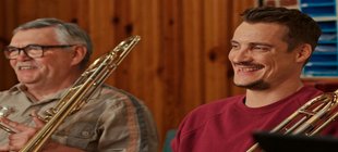 Zwei lachende Männer mit legerer Kleidung halten jeweils eine Posaune in der Hand.