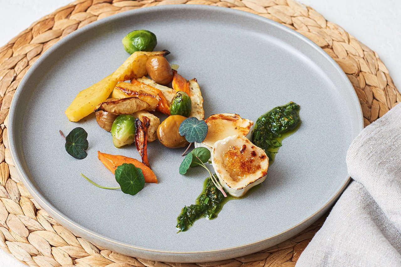 ein schlichter grauer Teller steht auf einem Bast-Tischset. In der Mitte ist buntes Wintergemüse angerichtet (Rosenkohl, Karotte, Maroni, Kartoffel), dazu gratinierter Ziegenkäse.