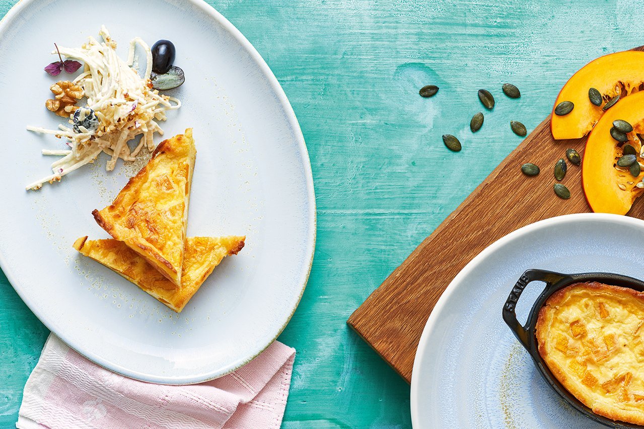 Auf einem weißen Teller, der auf einem kräftig türkisen Untergrund steht, sind zwei Scheiben Kürbisquiche mit Selleriesalat angerichtet. Am Rand steht eine weitere Ofen-Form mit Quiche auf einem Holzbrett