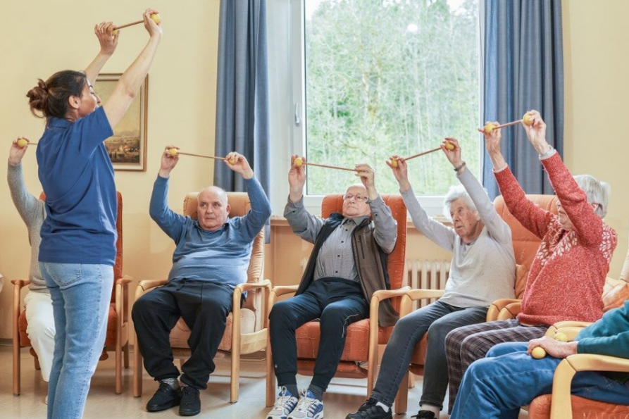 Eine Mitarbeiterin der Tagespflege zeigt einer Gruppe von Senior*innen eine Übung