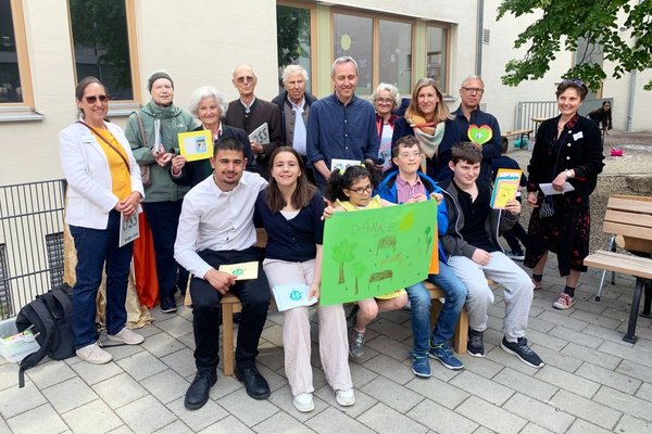 5 Kinder und Jugendliche sitzen auf einer Parkbank und halten ein Danke-Schild in die Kamera. Im Hintergrund stehen 10 Erwachsene, die Bänke gespendet haben