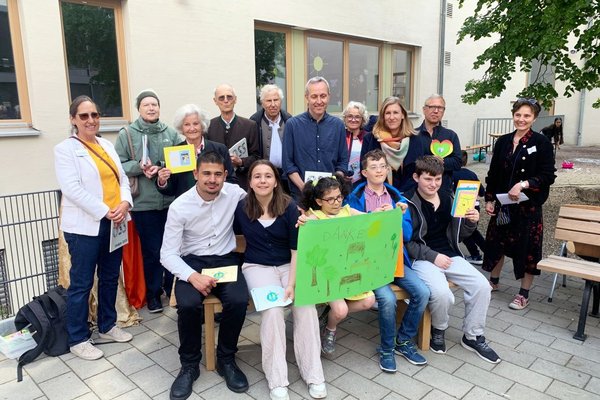 5 Kinder und Jugendliche sitzen auf einer Parkbank und halten ein Danke-Schild in die Kamera. Im Hintergrund stehen 10 Erwachsene, die Bänke gespendet haben