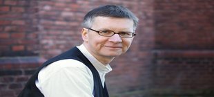 Ein Mann mit Brille in einem weißen Hemd und einer schwarzen Weste vor einer Ziegelmauer