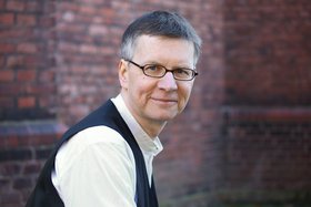 Ein Mann mit Brille in einem weißen Hemd und einer schwarzen Weste vor einer Ziegelmauer