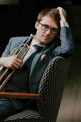 Junger Mann mit Brille, Anzug und Krawatte mit einer Trompete in der Hand