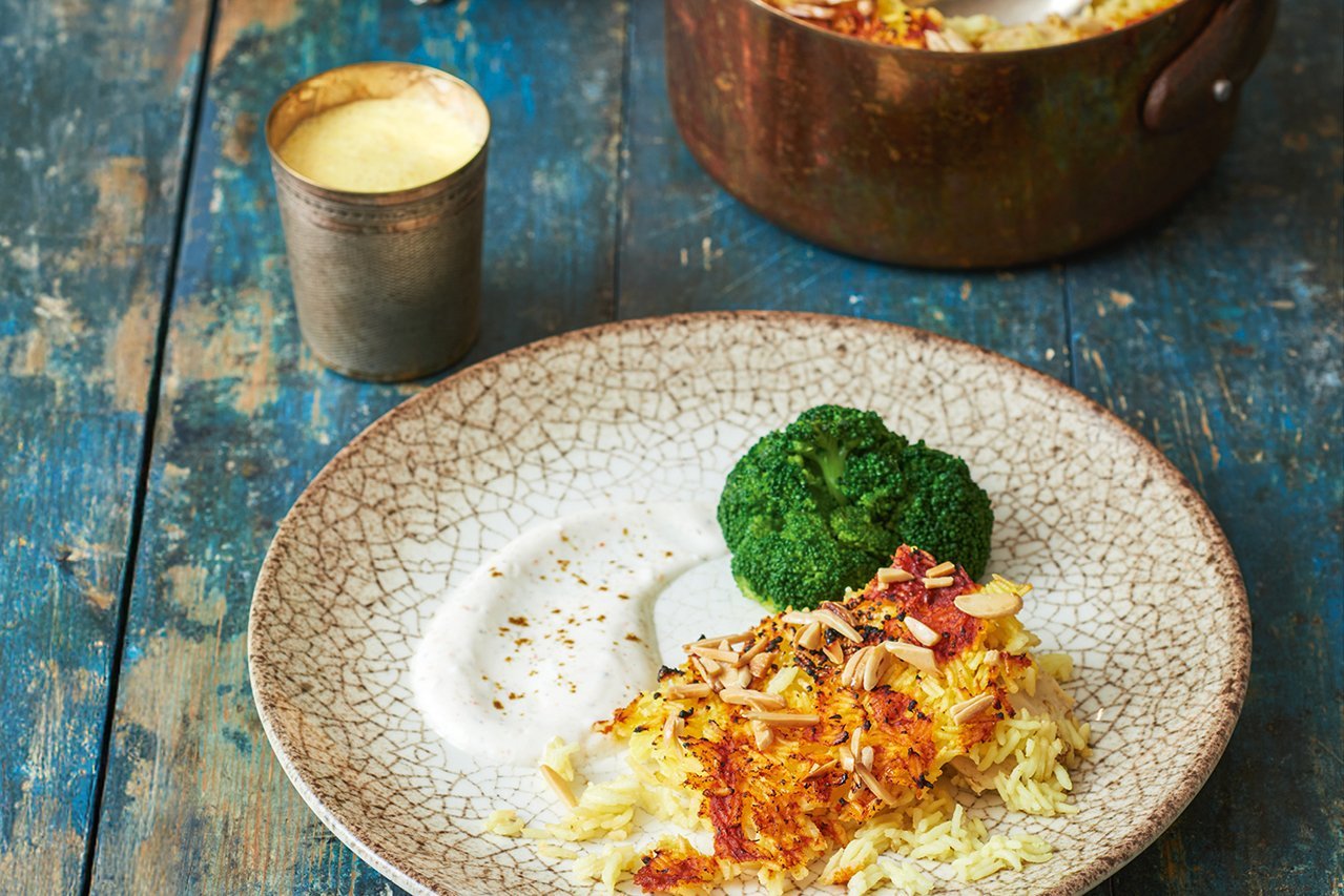 Ein braun-weißer Teller steht auf einem Shabby-Chic blauen Holzhintergrund. Hinter dem Teller steht ein Blechbecher mit gelbem Lassi. Auf dem Teller ist überbackener Reis mit Brokkoli und Joghurt angerichtet.